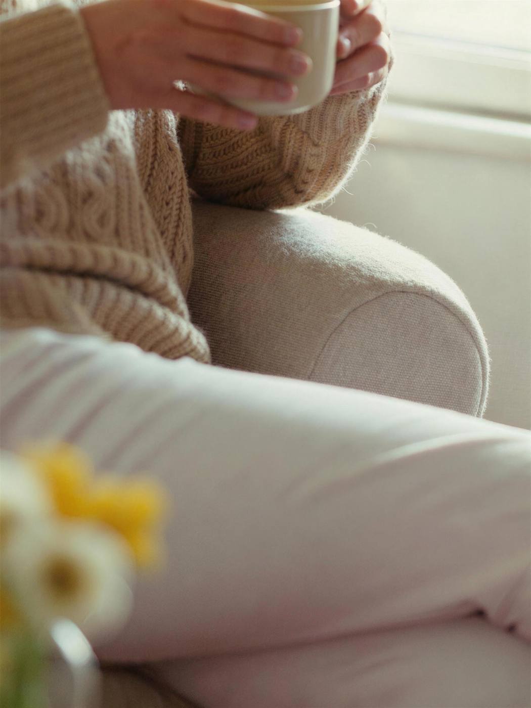 Eine Frau im warmen Strickpullover hält in einem Sessel eine Tasse Tee, ein ruhiger Moment für sich
